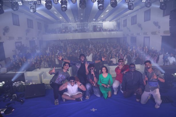 Group of performers posing on a blue stage, with a large cheering audience in a theater-like venue behind them.