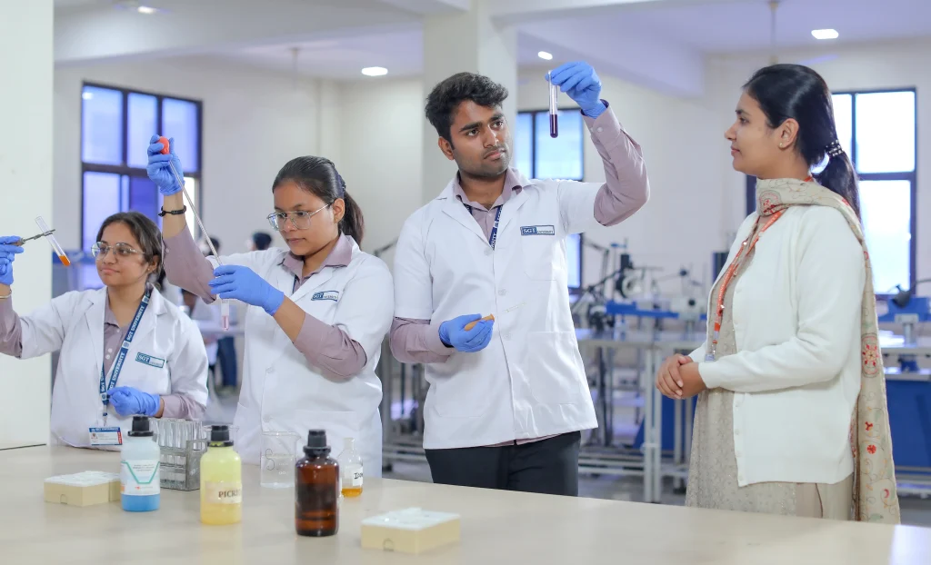 Group of students in lab coats and gloves handling test tubes while a woman observes in a laboratory setting.