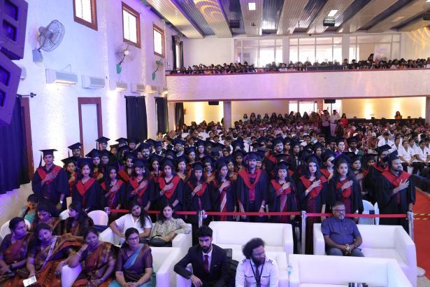 Graduates in caps and gowns stand in rows during a graduation ceremony in a large auditorium, with an audience seated behind them and on an upper level surface nearby.