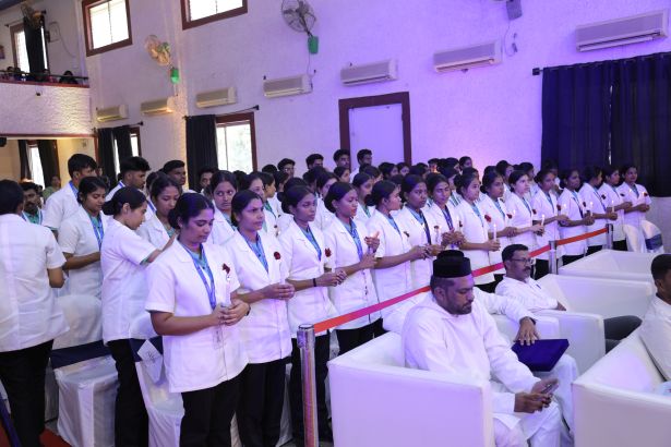 Nursing students in white uniforms stand in a line with hands clasped during a ceremony in a hall.