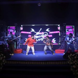 Five musicians perform on a lit stage with a bright LED backdrop, one in a red shirt center, two guitarists flanking, and floral arrangements at the front.