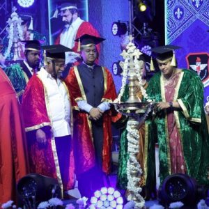 Group of graduates and officials in colorful gowns lighting a ceremonial lamp on stage during a graduation event.