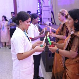 Nurse in a white uniform hands out colorful glow sticks to two women in sarees at a formal event, with others lining up in the background.